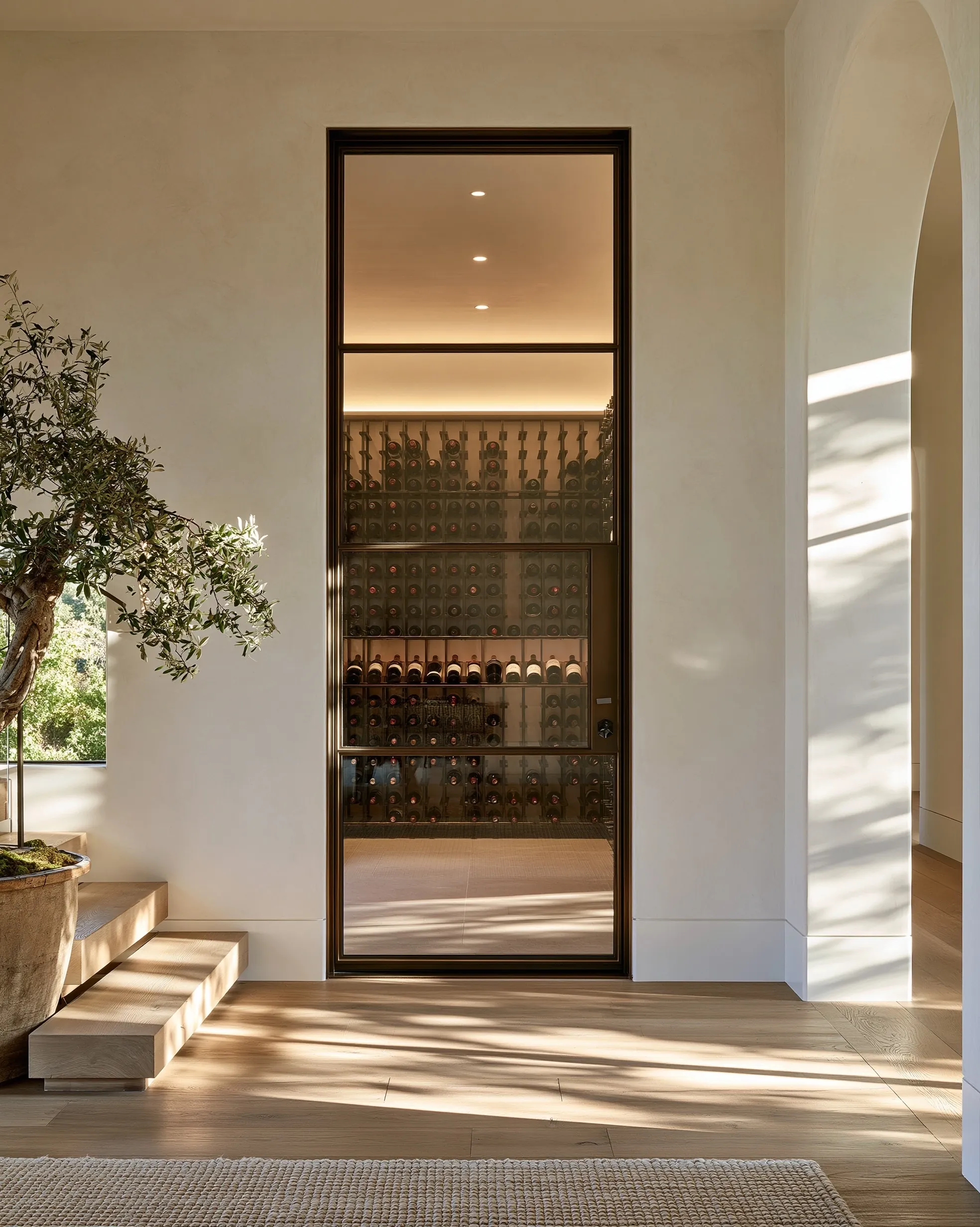 Tall glass cellar door in a slim bronze frame set into a plaster hallway wall, lit racking visible through it